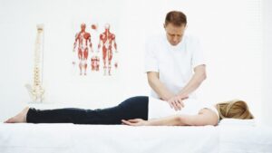 adult caucasian male performing chiropractic care for unknown age woman on a table, spinal cord model and chart displaying the human muscle system behind him