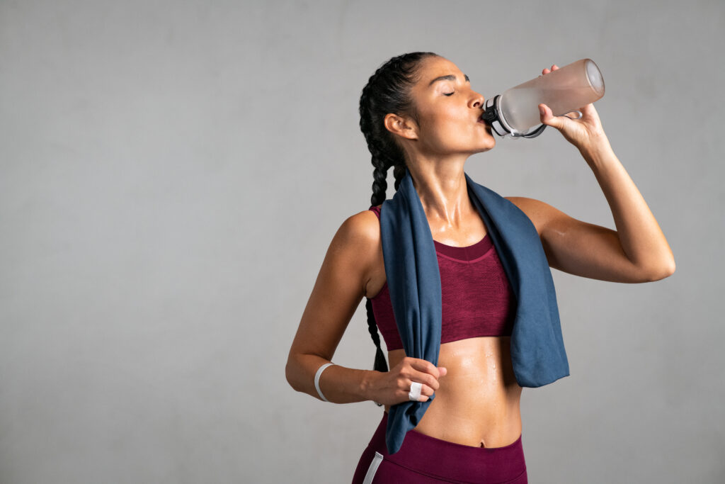 Fitness thirsty girl drinking water standing on gray background with copy space. Portrait of sweaty latin woman with towel around neck take a break after intense workout. Mid adult fit woman taking a break and drink from water bottle after gym workout isolated on grey wall.