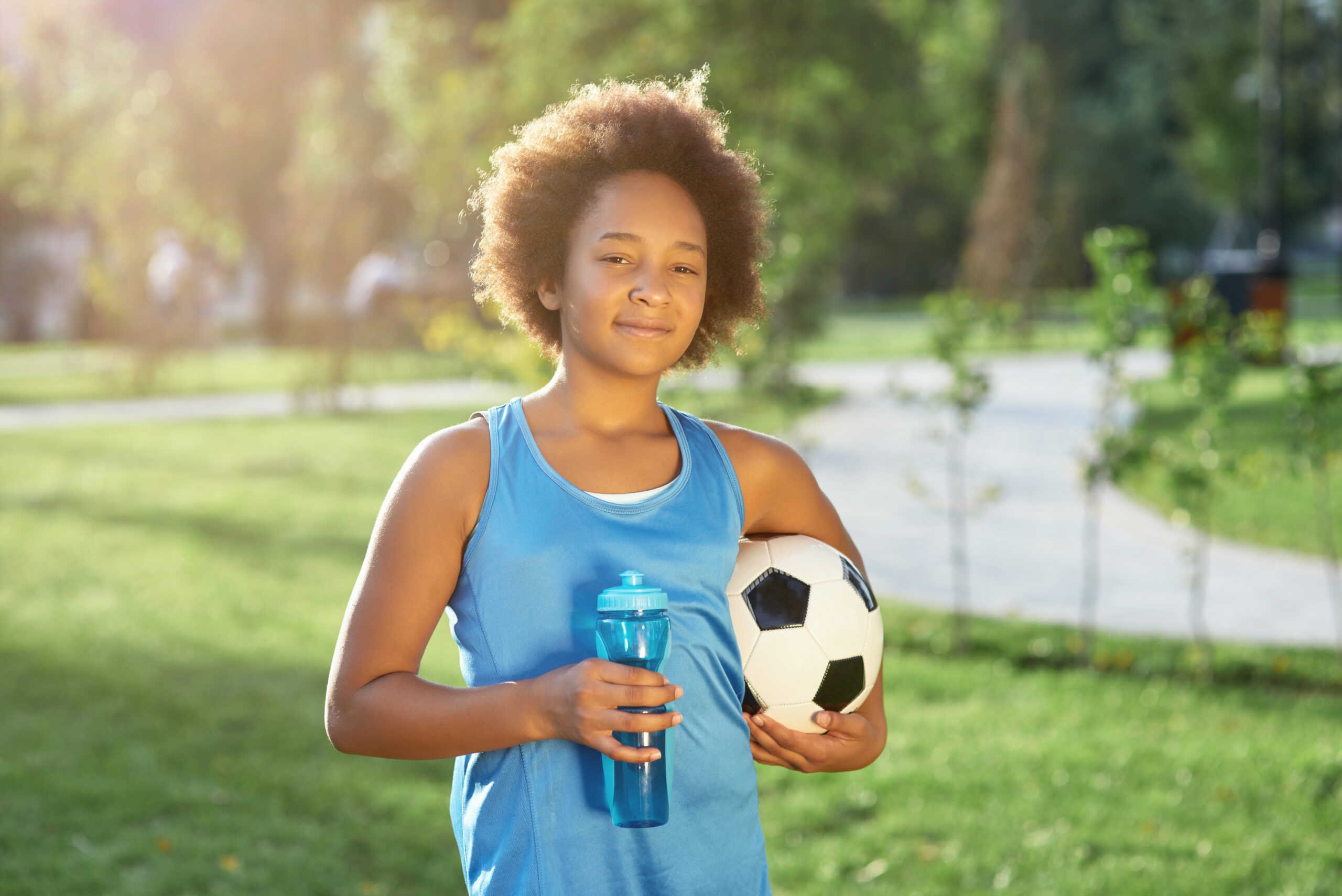 young african american female holding a water bottle and soccer ball, smiling at the camera against out of focus park background