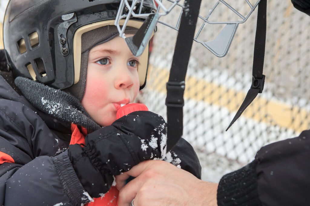 young boy dressed in winter gear and a hockey helmet drinking water given by adult male