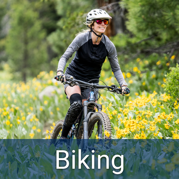 young woman wearing a helmet, sunglasses, and protective kneepads on a mountain bike, crossing a field of flowers, text below reads "Biking"