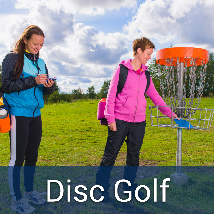 young adult female writes down score on a notepad while adult female retrieves frisbee from disc golf net on a cloudy but sunny day, bottom header: Disc Golf